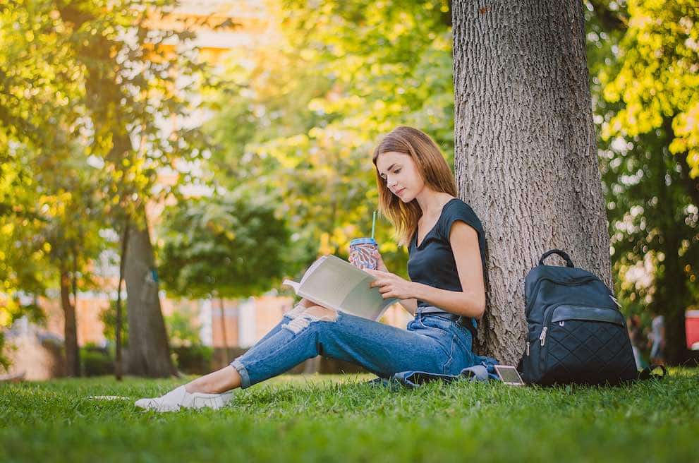 happy girl student sitting on the grass near the tree in the park and reading. Education concept